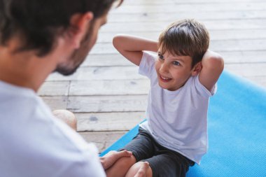 Genç bir adam evdeki oturma odasında egzersiz yaparak oğluna spor yaparken yardım ediyor..