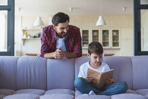 Счастливый маленький мальчик и его веселый папа читают книгу, сидя дома на диване в гостиной.