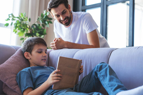 Happy little boy and his cheerful dad are reading a book sitting on the couch at home in the living room.