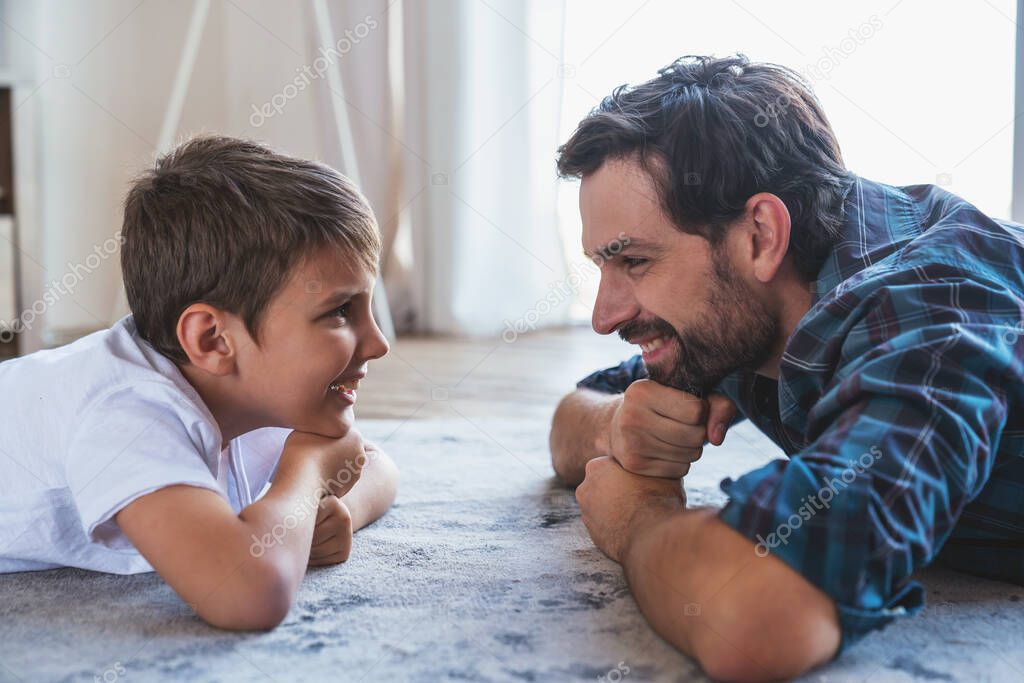 Portrait of a little boy and his dad lying on the floor and smiling looking at each other at home.