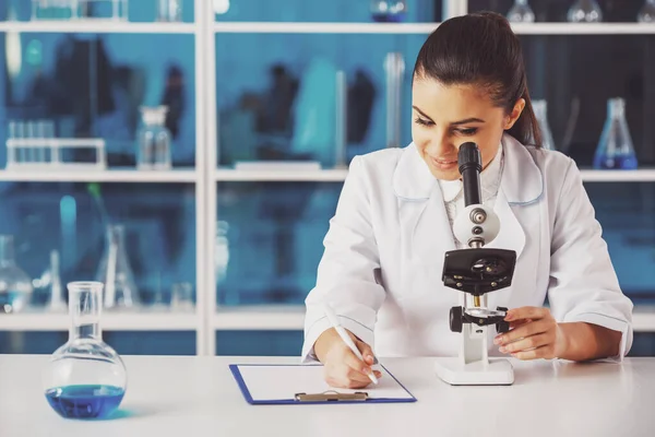 有吸引力的女科学家通过显微镜分析事物 并在实验室做笔记 — 图库照片