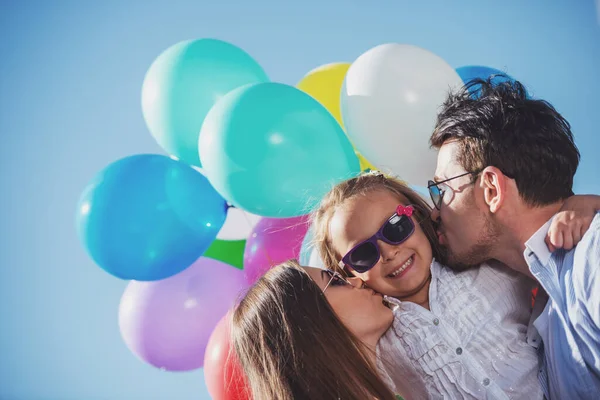 Young attractive parents with balloons kissing their little daughter on the cheek.