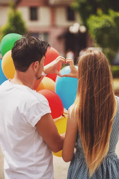 年轻可爱夫妇与气球塑造双手心符号后面观 — 图库照片