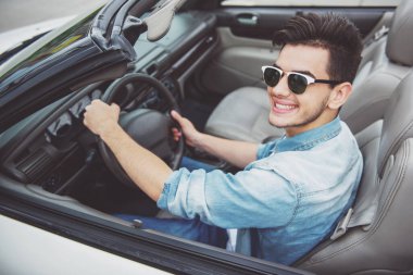 Yakışıklı adam cabriolet tarafından sürüş, kameraya bakıyor ve gülümseyen güneş gözlüğü.