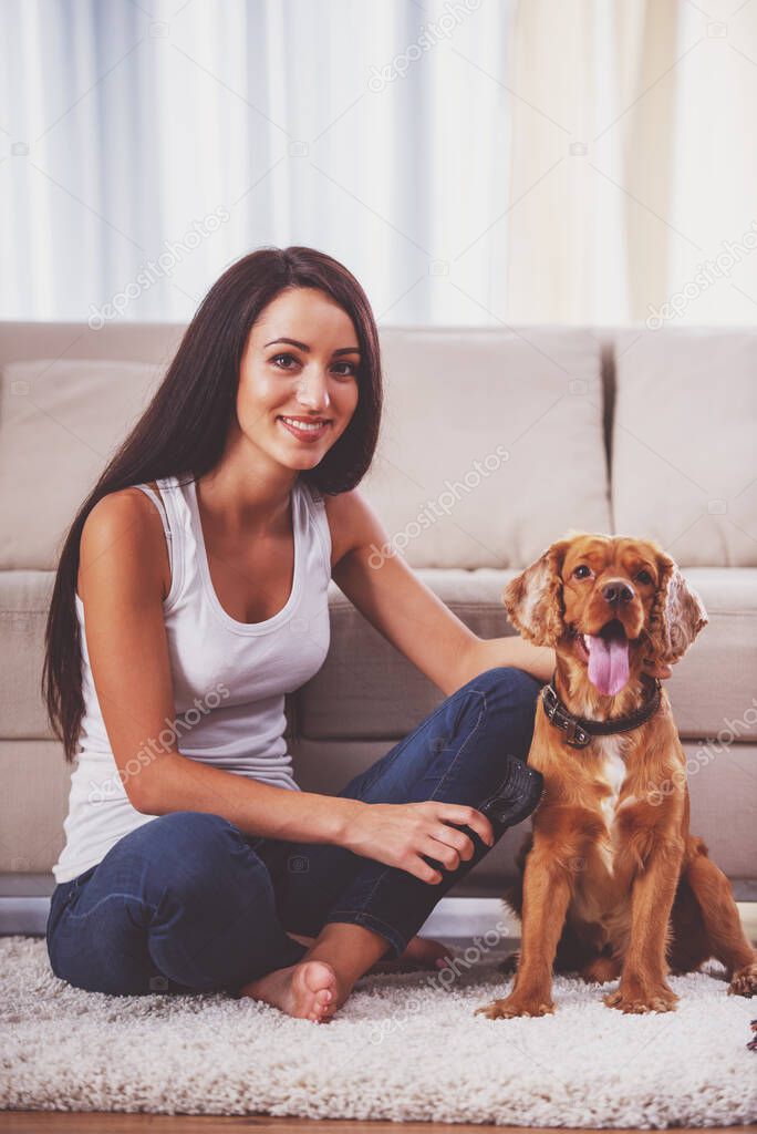 Smiling woman is sitting on the floor at her apartment with a dog.