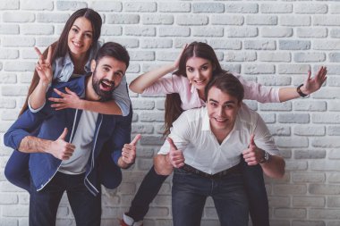 Bir grup mutlu arkadaş eğleniyor. Erkekler kızlara bir piggyback binmek veren, gülümseme, kameraya bakmak, elleriyle gesticulating, bir tuğla duvarın arka planında