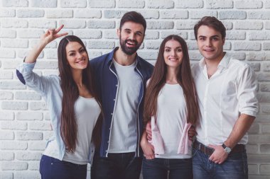 Günlük giysiler içinde dört genç güzel insanlar Grubu, stüdyoda poz, gülümseyerek, kameraya bakarak, bir tuğla duvara karşı