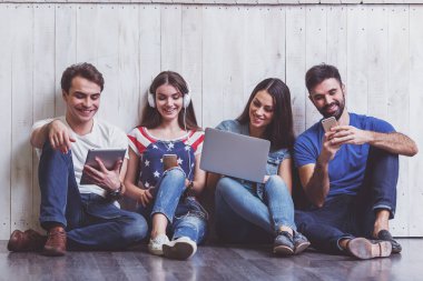 Kat bir dizüstü bilgisayar, Tablet Pc, akıllı telefonlar, müzik dinlerken kulaklık kullanarak üzerinde oturan çekici genç insanlar grup gülümseyen