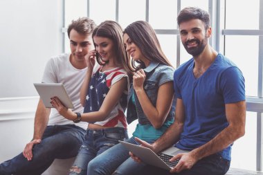 Dört genç çekici insanların dizüstü ve tablet Pc, pencere pervazına üzerinde oturan kullanarak gülümseyen