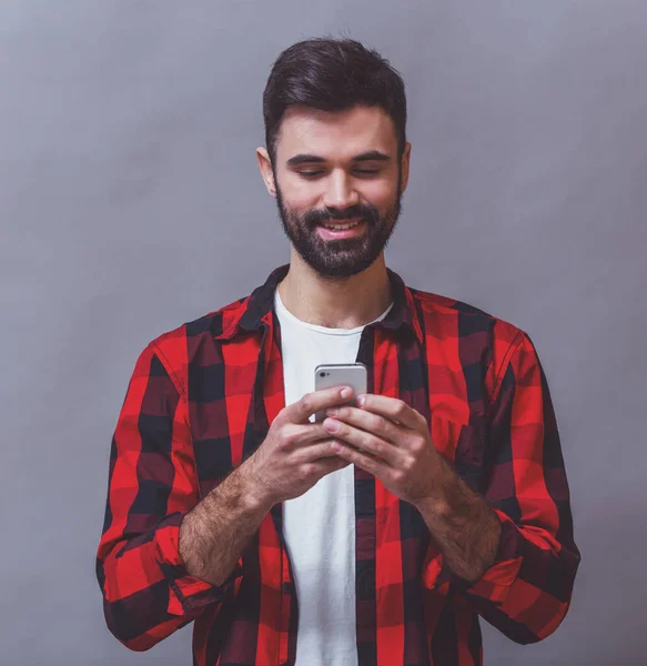 灰色の背景に笑みを浮かべて スマート フォンを使用して 市松模様のシャツを着た 髭の若い男の肖像 — ストック写真