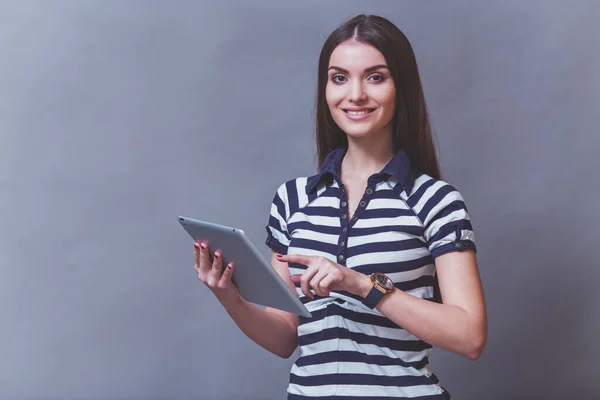 一个穿着条纹衬衫的年轻美女的肖像 使用平板电脑 面带微笑 看着相机 在灰色背景 — 图库照片