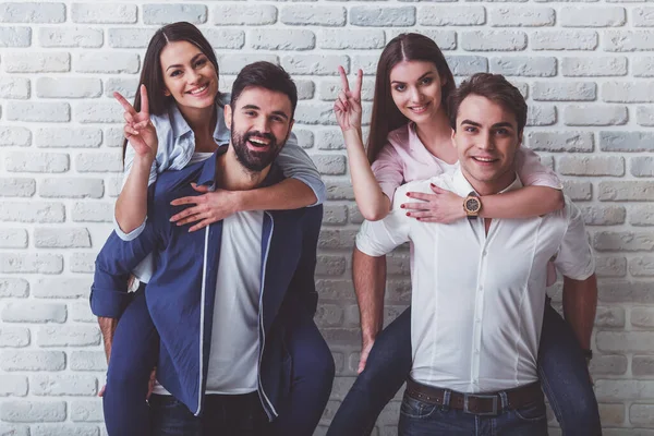 Bir grup mutlu arkadaş eğleniyor. Erkekler kızlara bir piggyback binmek veren, gülümseme, kameraya bakmak, elleriyle gesticulating, bir tuğla duvarın arka planında