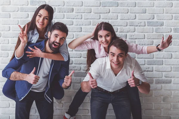 Bir grup mutlu arkadaş eğleniyor. Erkekler kızlara bir piggyback binmek veren, gülümseme, kameraya bakmak, elleriyle gesticulating, bir tuğla duvarın arka planında