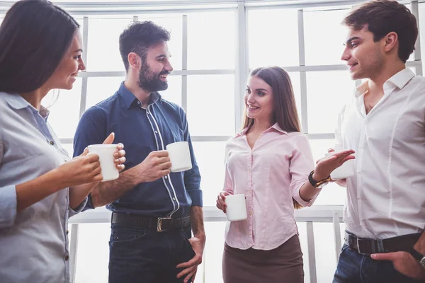 Coffee break chat. Group of attractive business people, standing next to each other, holding a cups, smiling standing at the window