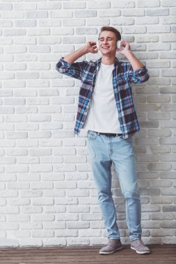 Giyinmiş genç yakışıklı adam moda giysiler içinde bir tuğla duvara cep telefonunda kulaklık ile müzik dinlemek