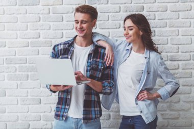 Çekici bir genç çift, ayakta ve bir dizüstü bilgisayar, bir gülümseme ile ekrana bakarak bir tuğla duvar bir arka plan üzerinde tutan
