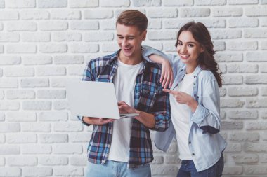 Çekici bir genç çift, ayakta ve bir dizüstü bilgisayar, bir gülümseme ile ekrana bakarak bir tuğla duvar bir arka plan üzerinde tutan