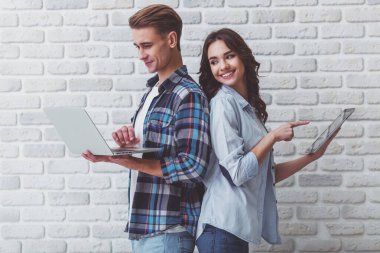 Bir dizüstü bilgisayar ve bir tablet bir tuğla duvar arka plan üzerinde genç güzel çift dur