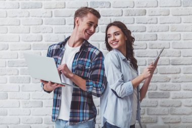 Bir dizüstü bilgisayar ve bir tablet bir tuğla duvar arka plan üzerinde genç güzel çift dur