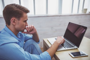 Yakışıklı delikanlı mavi tişört ve kot pantolon, düşünme kullanır tablo sonraki smartphone bir laptop yerde evde oturuyor