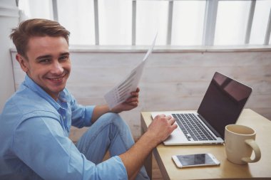 Yakışıklı delikanlı mavi tişört ve kot pantolon, masada bir dizüstü bilgisayar kullanarak, sonraki katta evde oturan bir grafik tutarak bir smartphone ve bir fincan.