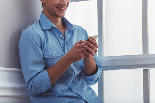 自宅の窓辺に座っているスマート フォンを使用して青いシャツと光のジーンズで美しい若い男 — ストック写真
