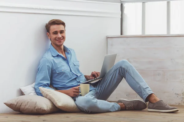 年轻英俊的男人穿着蓝色衬衫和牛仔裤 与一杯在手 使用手提电脑 坐在地上 垫上接下来的是图形 坐在家里的地板上 — 图库照片
