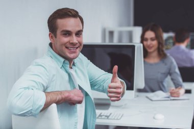 Günlük giysiler içinde genç ve çekici bir iş adamı tamam işareti gösteriyor, gülümsüyor ve arka planda ofiste çalışan insanlara bakıyor.