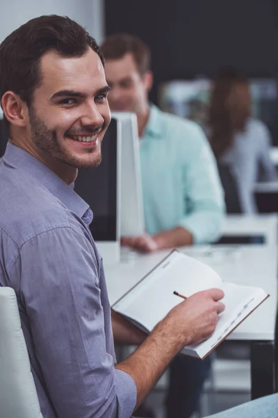 カジュアルな服を着た若い魅力的なビジネスマンは オフィスで働く人々を背景に カメラを見て笑顔でメモを作ります — ストック写真