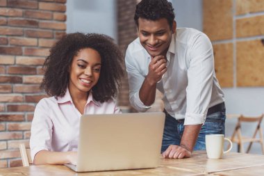 Genç ve çekici Afro-Amerikan iş çifti laptopu kullanıyor, kafede çalışırken konuşuyor ve gülümsüyor.