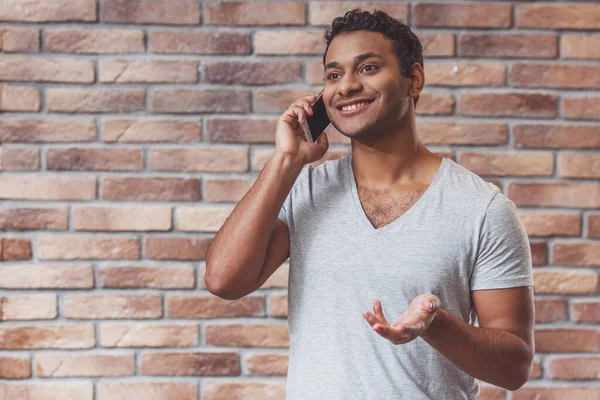 若いですハンサムなアフリカ系アメリカ人の男は レンガの壁に向かって立っている間 電話で話して笑顔 — ストック写真