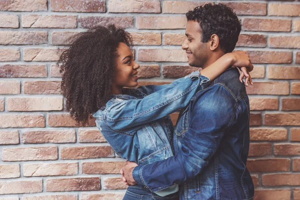 侧视图的年轻有吸引力的非洲裔美国夫妇拥抱 看着相机和微笑 而站在砖墙 — 图库照片