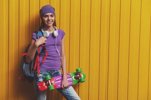 可爱少女穿着 恤和帽地位与学校背包 耳机上橙色的背景下 举行一个滑板 看在相机和微笑 — 图库照片
