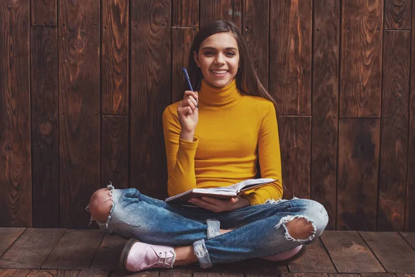 可爱的少女拿着一支钢笔和一本笔记本 看在相机 微笑着坐在木制的背景下 — 图库照片