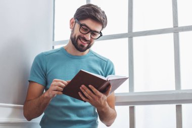 Yakışıklı adam rahat kıyafetler ve bir kitap okuma ve pencerenin yanında otururken gülümseyen gözlük