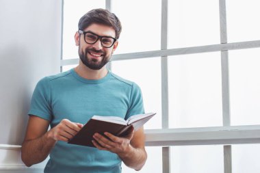 Yakışıklı adam rahat kıyafetler ve bir kitap okuma, kamera seyir ve pencerenin yanında otururken gülümseyen gözlük
