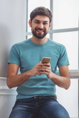 Bir cep telefonu kullanarak, kamera seyir ve pencerenin yanında otururken gülümseyen rahat kıyafetler içinde yakışıklı adam