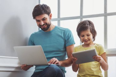 Bir dizüstü bilgisayar ve pencerenin yanında otururken tablet kullanma şirin küçük oğlu kullanarak rahat kıyafetler içinde yakışıklı genç Baba