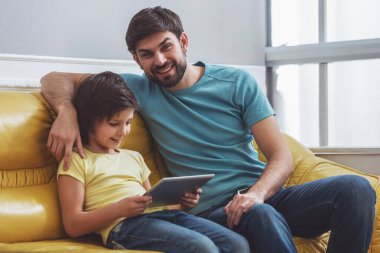 Kamera seyir ve sevimli küçük oğlu, yakındaki bir kanepede otururken gülümseyen rahat kıyafetler içinde yakışıklı genç Baba kim tablet kullanma