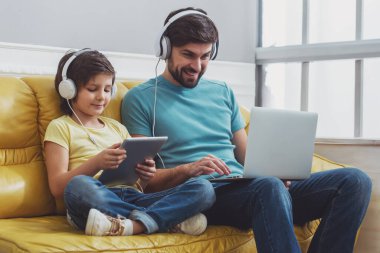Yakışıklı genç Baba bir dizüstü ve tablet odasında bir kanepede otururken kullanma şirin küçük oğlu kullanarak rahat kıyafetler içinde. İkimiz de kulaklık.