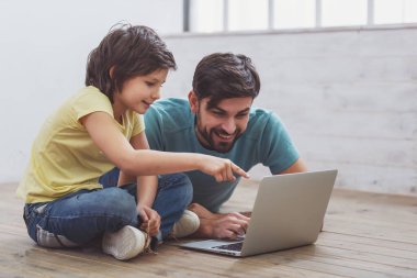 Yakışıklı genç Baba rahat kıyafetler ve odada bir ahşap zemin üzerinde yalan bir dizüstü bilgisayar kullanarak sevimli küçük oğlu