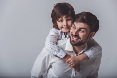 Portre sevimli oğlu sırtında taşıyan ve gülümseyen yakışıklı bir baba. İkimiz de gri bir arka plan ayakta beyaz klasik gömlek.