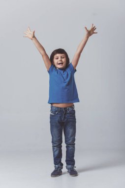 Kamera seyir ve gri arka plan üzerinde dururken gülümseyen gösteren mutluluktan ellerini, gergin bir mavi t-shirt şirin küçük çocuk