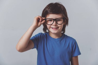 Mavi bir tişört ve kamera seyir ve gri arka plan üzerinde dururken gülümseyen gözlük şirin küçük bir çocuk portresi