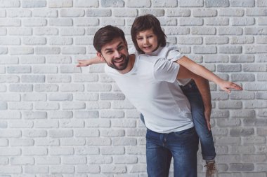 Portre sevimli oğlu sırtında taşıyan bir yakışıklı baba. İkimiz de gülümseyerek, beyaz duvara karşı ayakta beyaz t-shirt.