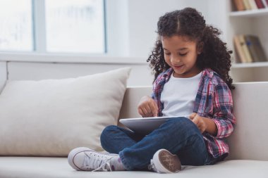 Şirin Afro-Amerikan kız tablet kullanma ve odada bir kanepede otururken gülümseyen rahat kıyafetler.