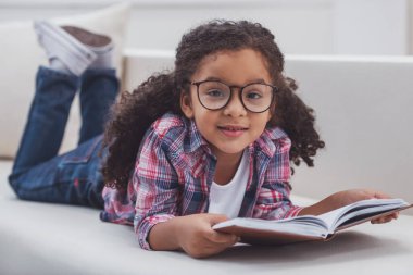 Sevimli küçük Afro-Amerikan kız rahat kıyafetler ve bir kitap okuma, kameraya bakıyor ve gülümseyen bir kanepe odasında yatarken gözlük.