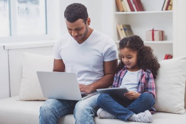 Sevimli küçük Afro-Amerikan kız rahat kıyafetler ve yakışıklı babası odasında bir kanepede otururken bir tablet ve bir dizüstü bilgisayar kullanarak.