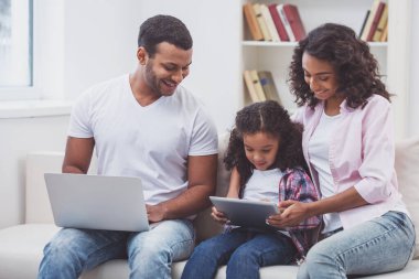 Sevimli küçük Afro-Amerikan kız ve odada bir kanepede oturan güzel genç ailesi. Bir dizüstü bilgisayar, anne ve kızı bir tablet kullanarak kullanarak baba.