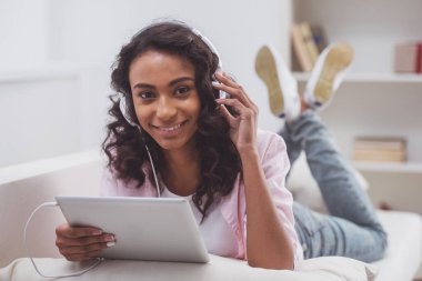 Müzik, kameraya bakarak ve bir kanepe odasında yatarken gülümseyen genç güzel Afro-Amerikan kadını, bir dizüstü bilgisayar kullanarak kulaklık.